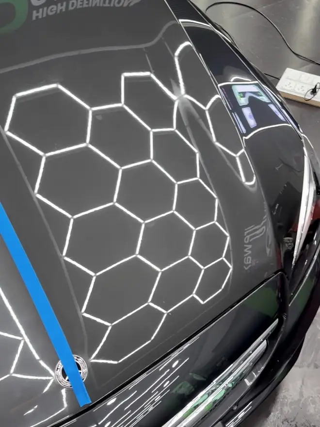 Hexagon light grid reflected across a dark bonnet with masking tape at one edge